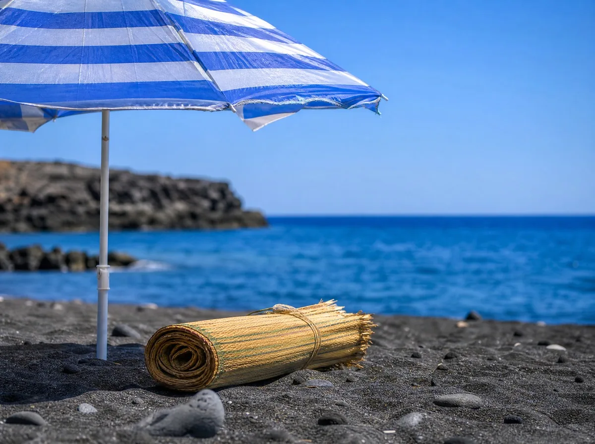 A blue-striped beach umbrella planted in dark sand, a rolled mat beside it and the open sea behind
