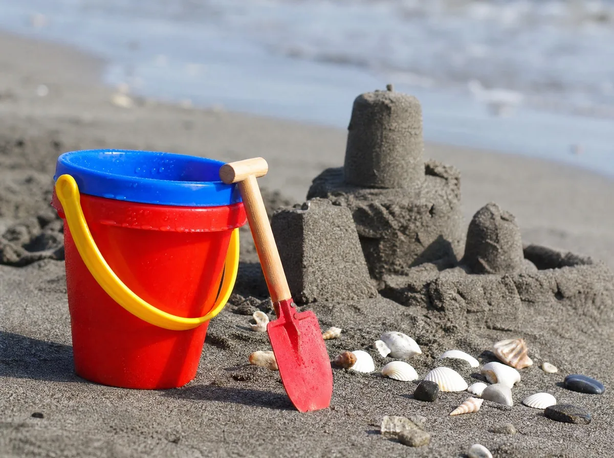 A bucket and spade left on dark sand beside a sandcastle and a scatter of shells